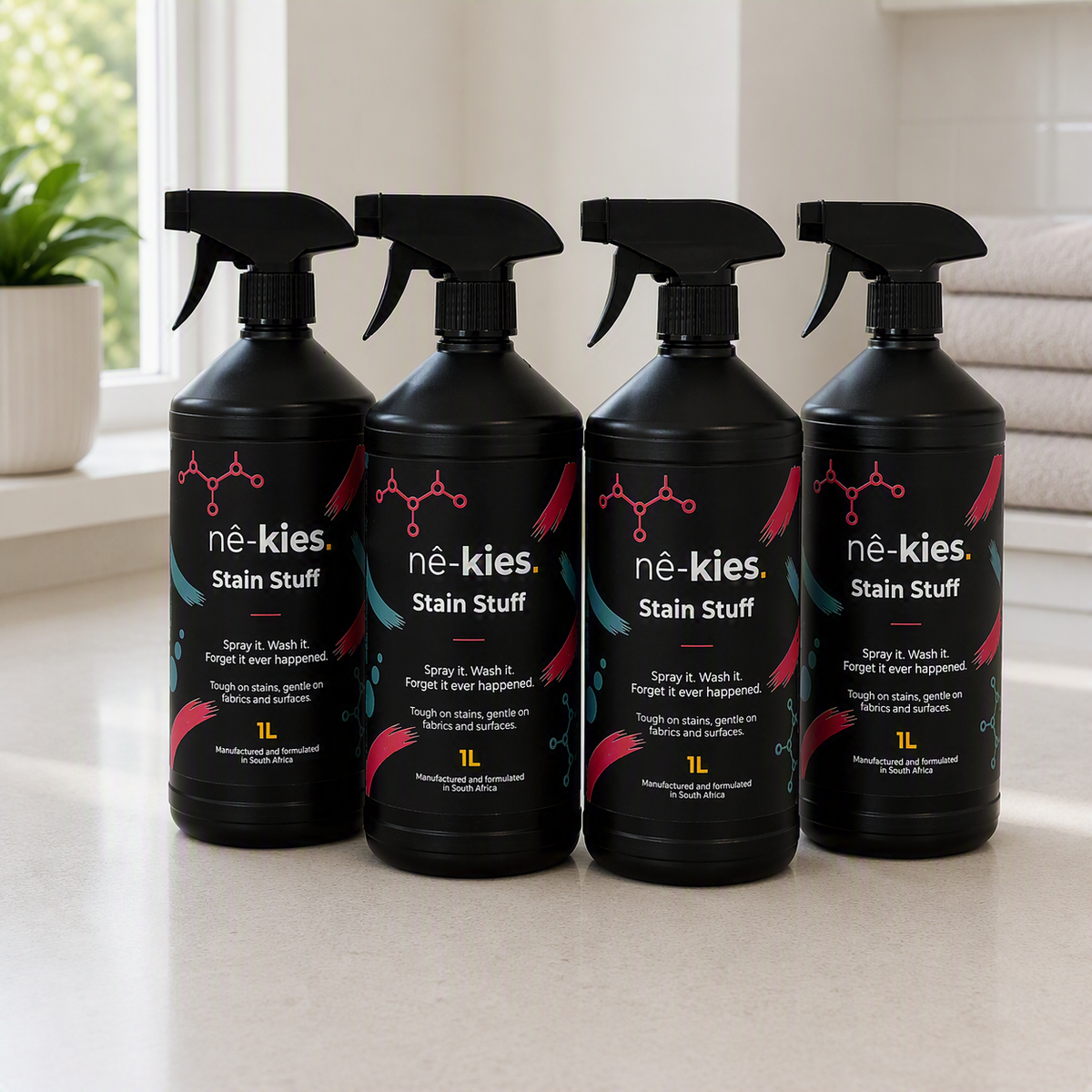 Four black spray bottles of Nê-kies Stain Stuff arranged side by side on a light countertop near a window.