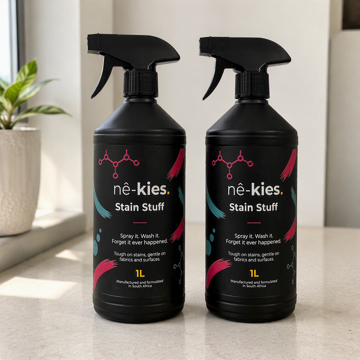 Two black spray bottles of Nê-kies Stain Stuff placed side by side on a light countertop, with a blurred indoor background and a potted plant near the window.