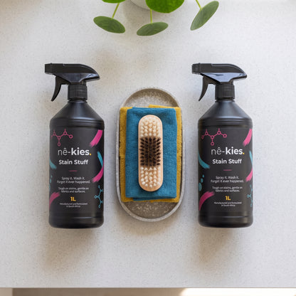 Two 1L bottles of né-kies Stain Stuff placed on either side of a small brush resting on a blue and yellow cloth, arranged on a light stone surface with green leaves at the top.