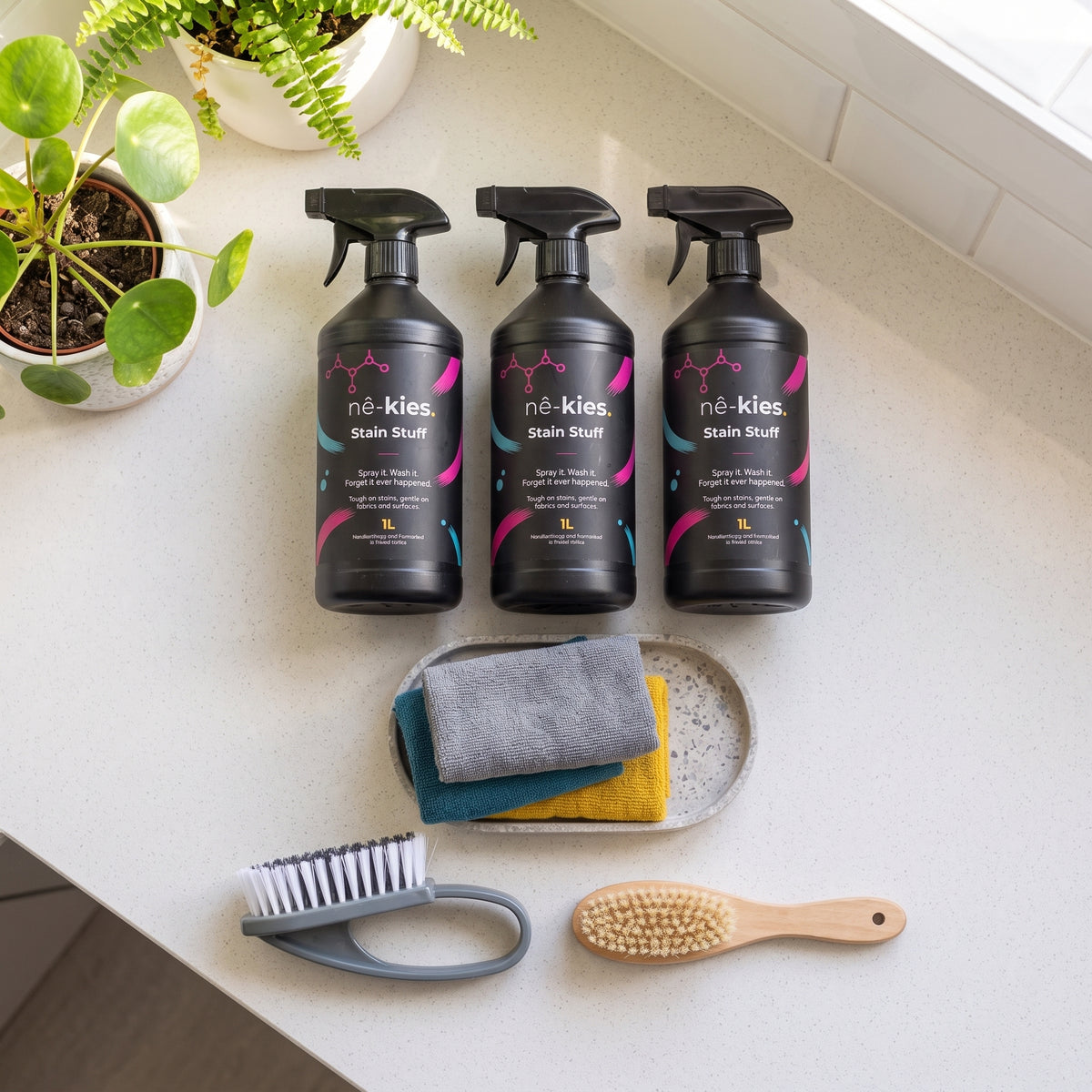 Three black Né-Kies Stain Stuffer bottles arranged on a light countertop with cleaning accessories below, including a small tray with folded cloths, a scrubber, and a wooden brush, alongside potted plants near a window.