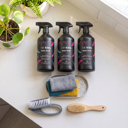 Three black Né-Kies Stain Stuffer bottles arranged on a light countertop with cleaning accessories below, including a small tray with folded cloths, a scrubber, and a wooden brush, alongside potted plants near a window.