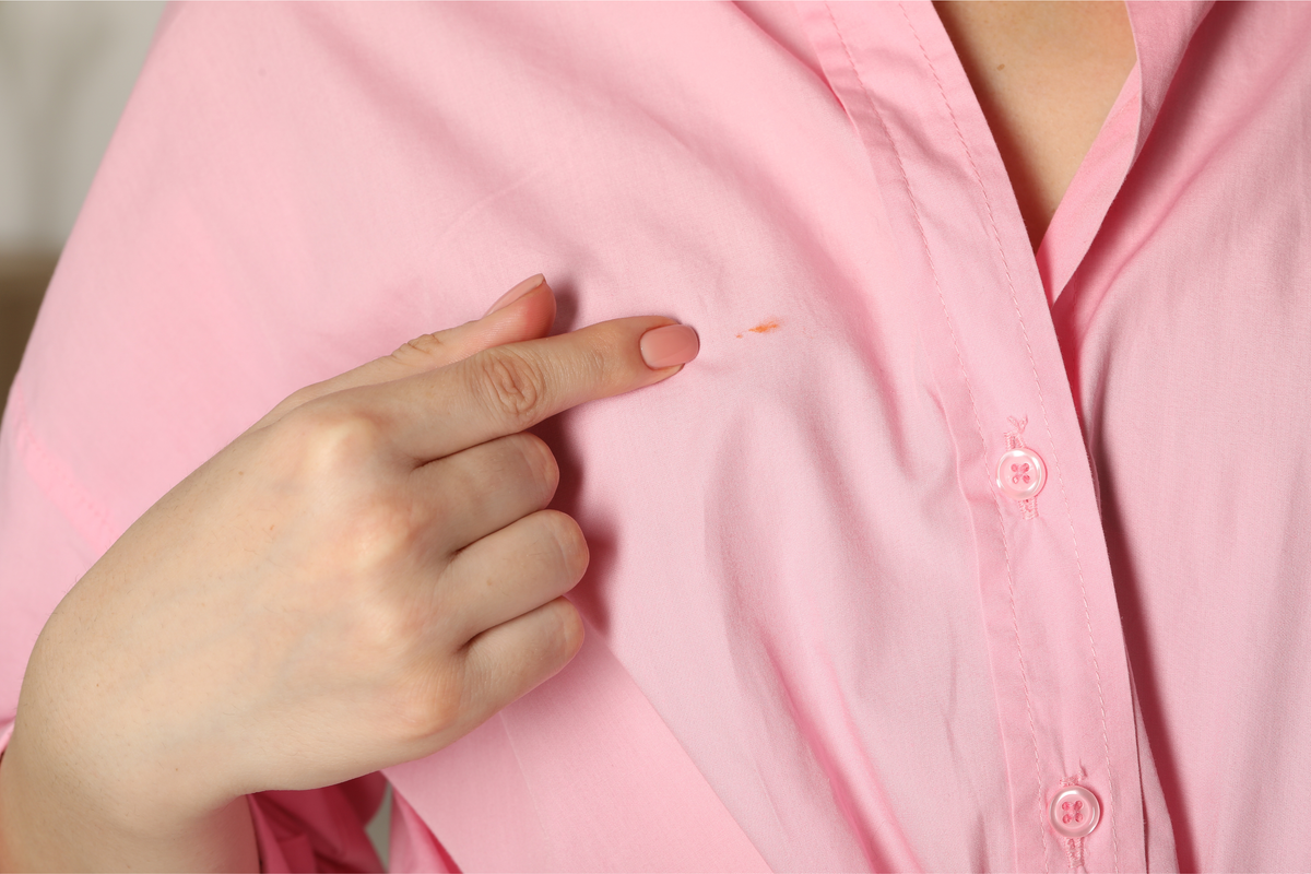 Person wearing a pink shirt with a small mark on it, pointing to the mark with their finger.